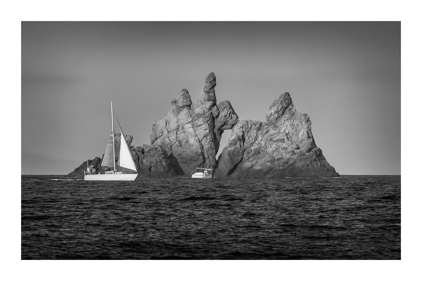 Voilier longeant un massif rocheux percé d’une arche en Méditerranée, lumière rasante de fin de journée, noir et blanc avec bordure